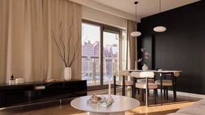 Ein modernes Wohnzimmer mit einem runden Couchtisch, einem Essbereich mit weißem Tisch und Stühlen, einer schwarzen Wand und einem großen Fenster mit Blick auf Gebäude.