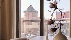 Ein Zimmer mit großem Fenster, einer Vase mit getrockneten Blumen und Blick auf einen historischen Turm.
