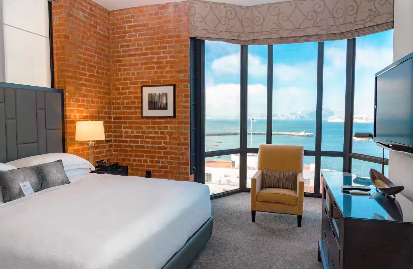 A hotel room with a brick wall, a bed, a chair, a desk with a TV, and a large window offering a view of the sea.