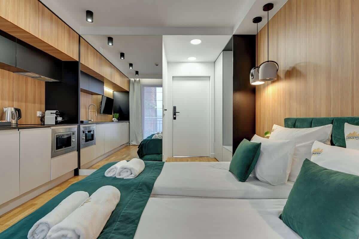 Ein modernes Hotelzimmer mit Küchenzeile, einem Bett mit grüner Bettwäsche und Blick auf den Flur.