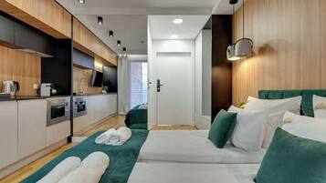 Ein modernes Hotelzimmer mit Küchenzeile, einem Bett mit grüner Bettwäsche und Blick auf den Flur.