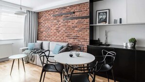 Un espace de vie moderne avec un mur d’accent en briques, un canapé avec des coussins, une table de salle à manger ronde avec des chaises, un coin cuisine doté d’un comptoir noir et un luminaire suspendu.