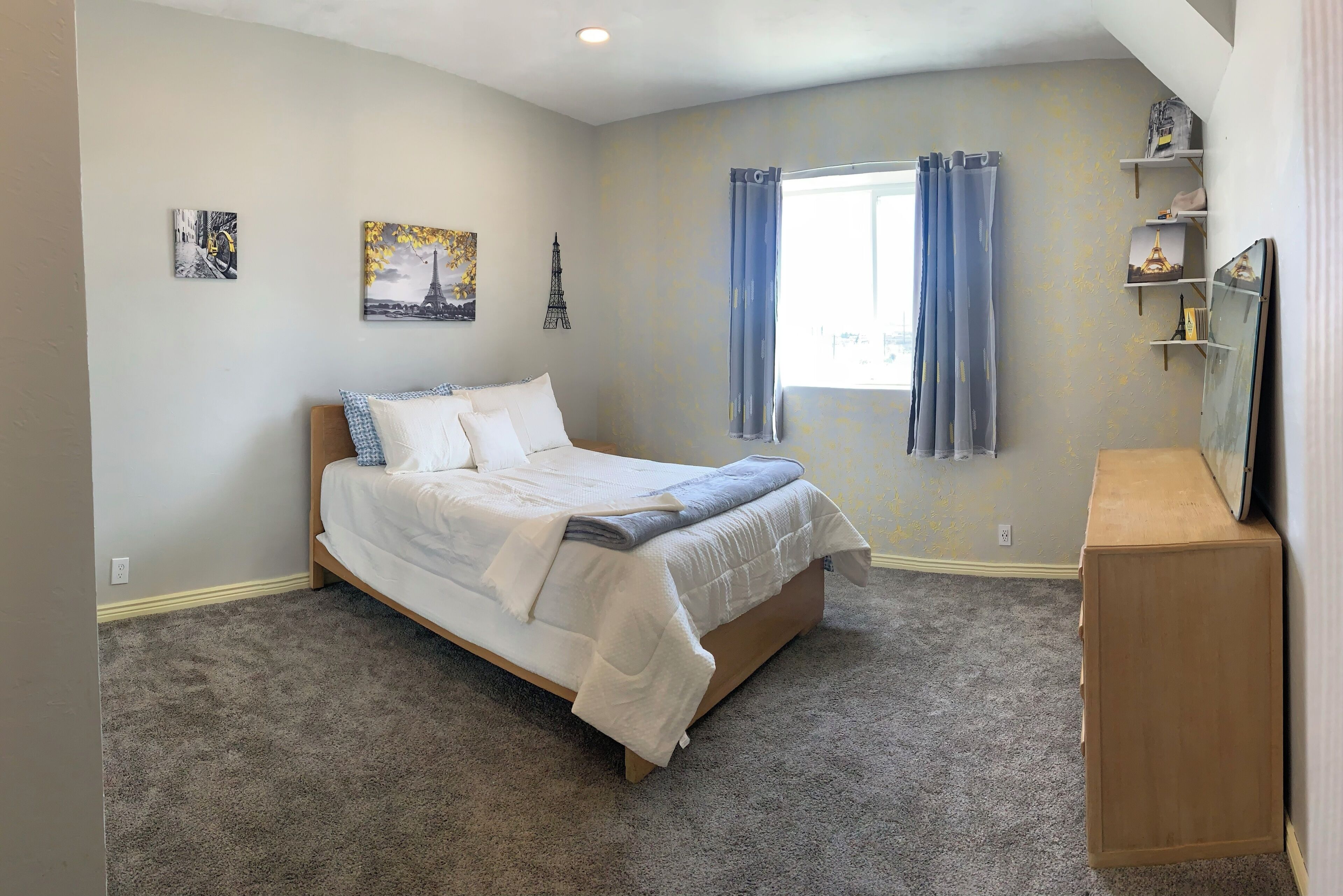 Une chambre à coucher avec un lit, une fenêtre avec des rideaux, une étagère avec des objets décoratifs et un mur orné d’un tableau.
