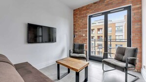 A modern living room with a flat-screen TV, a wooden coffee table, two grey armchairs, and a brick accent wall. - Aura III 36 Deluxe | River View | City Center (Gdańsk)