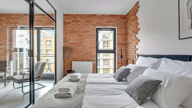 A modern hotel room with a large bed, a view of the city, and a brick wall. - Aura III 36 Deluxe | River View | City Center (Gdańsk)