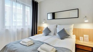 Une chambre d’hôtel avec un grand lit, une table de chevet avec une lampe, un miroir au-dessus du lit et des rideaux légers à la fenêtre.