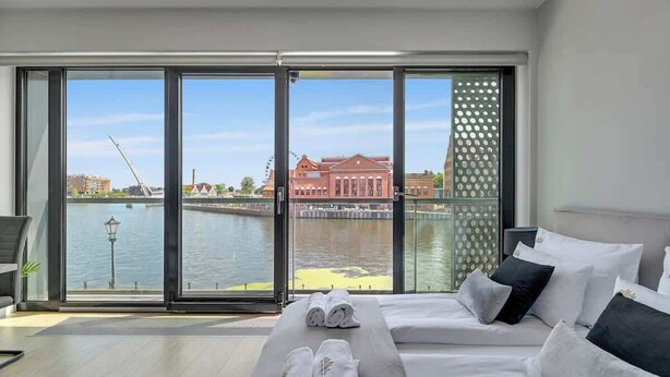 Habitación de hotel con un ventanal grande, una cama con ropa blanca y vistas a un cuerpo de agua y edificios.