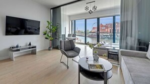 Una sala de estar moderna con un televisor de pantalla plana, un sofá gris, una mesa de centro redonda y un amplio ventanal con vistas a un paisaje urbano.