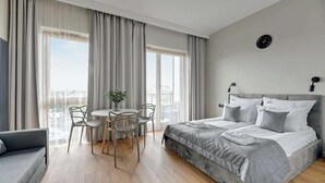 Moderni hotellihuone, jossa on suuri sänky, pyöreä ruokapöytä tuoleineen, pieni ruukkukasvi ja näkymä kaupunkiin.