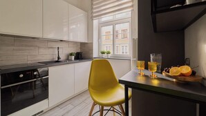 Une cuisine moderne avec des meubles blancs, une chaise jaune, une fenêtre avec des stores et un saladier rempli d’oranges et de raisins.
