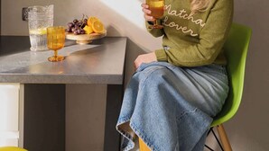 Une personne est assise à une table devant une boisson, une assiette de fruits et une étagère remplie d’ustensiles de cuisine.