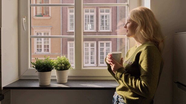 Une personne se tient près d’une fenêtre, avec deux plantes en pot, et tient une tasse à la main.