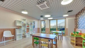 Une salle de classe avec des tables et des chaises en bois, des étagères blanches et une vue par les fenêtres.