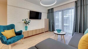 Een moderne woonkamer met een flatscreen televisie, een blauwe fauteuil, een grijze bank, een klein rond tafeltje en een groot raam met gordijnen.