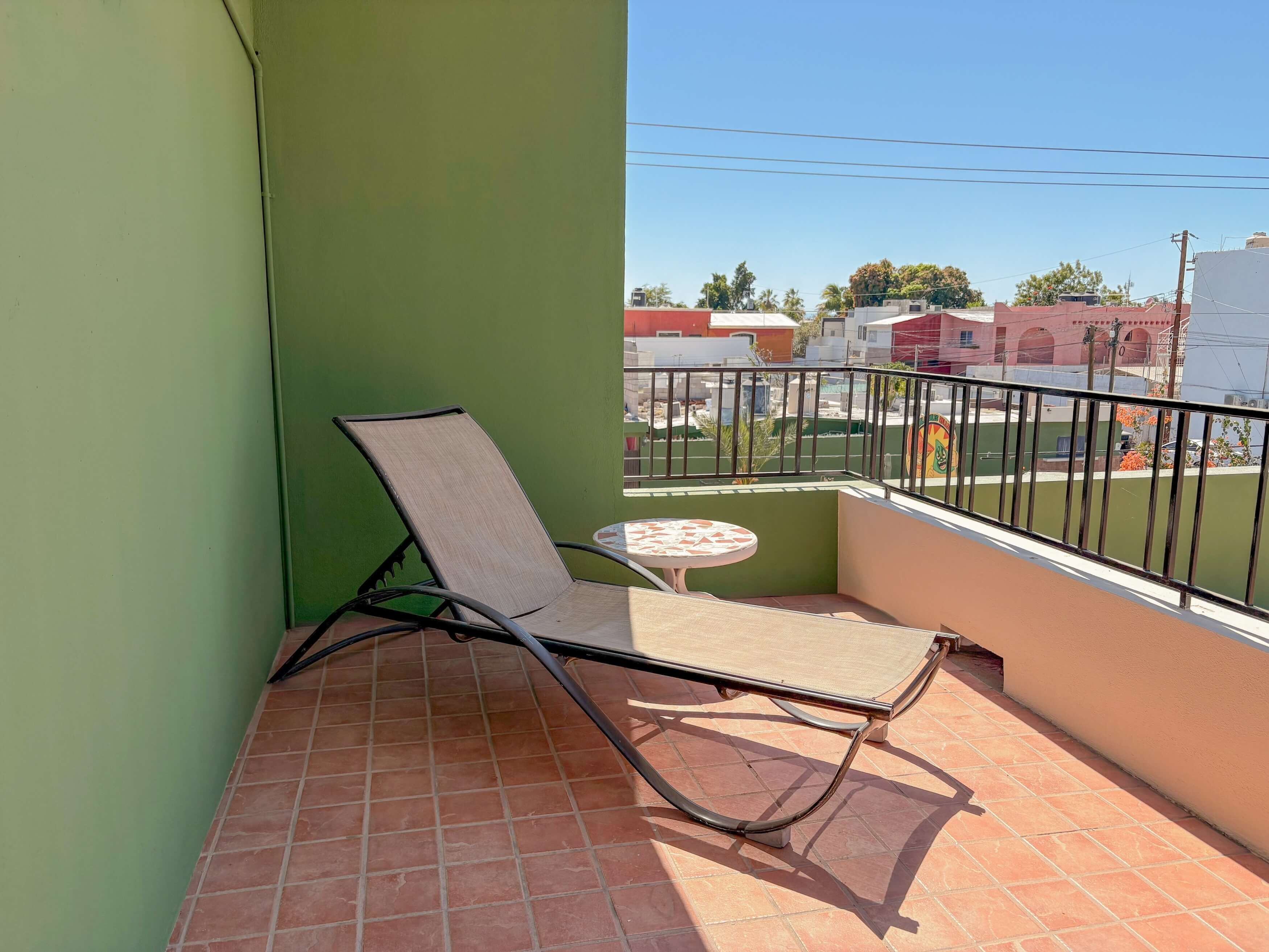 Un balcón con una sola silla y una mesita, un muro verde, y vista de edificios al fondo.