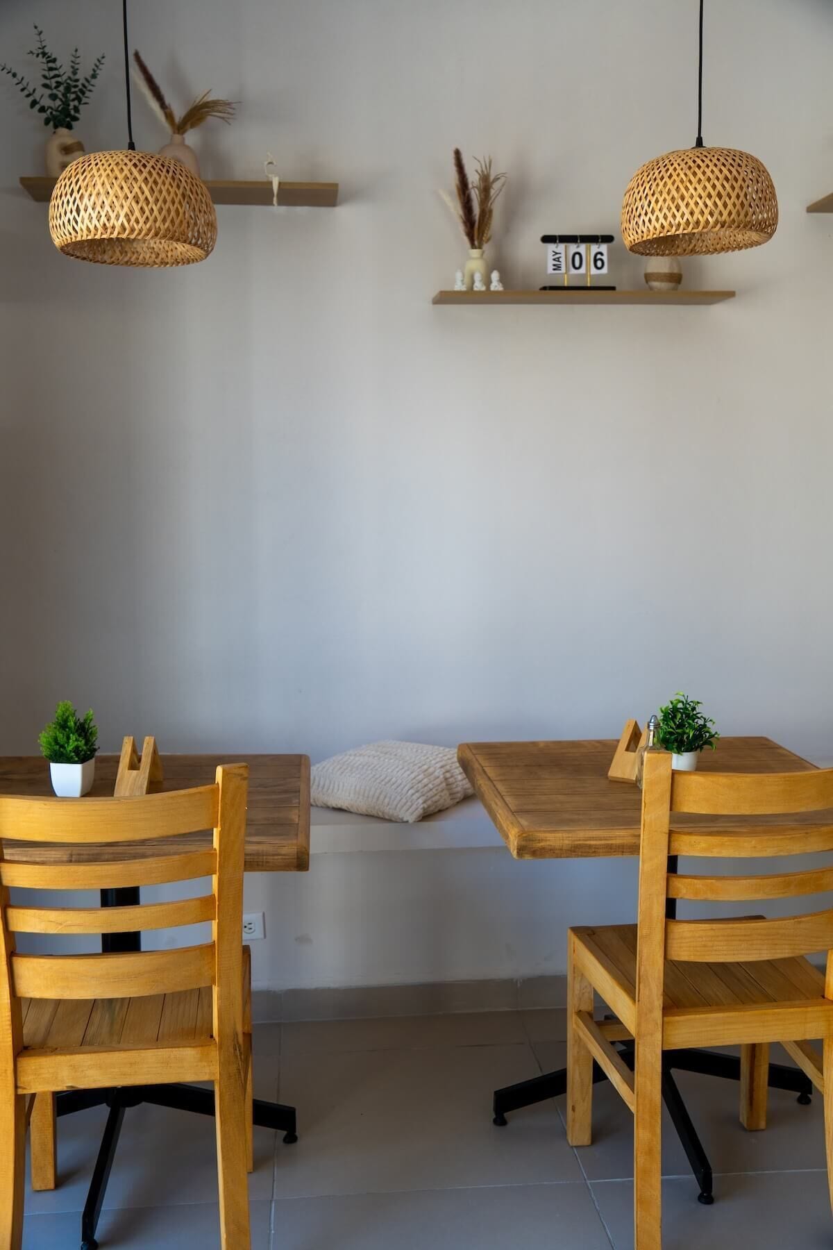 Uma sala de jantar com uma mesa e cadeiras de madeira, duas luminárias de rattan suspensas e uma estante fixada na parede com objetos decorativos.