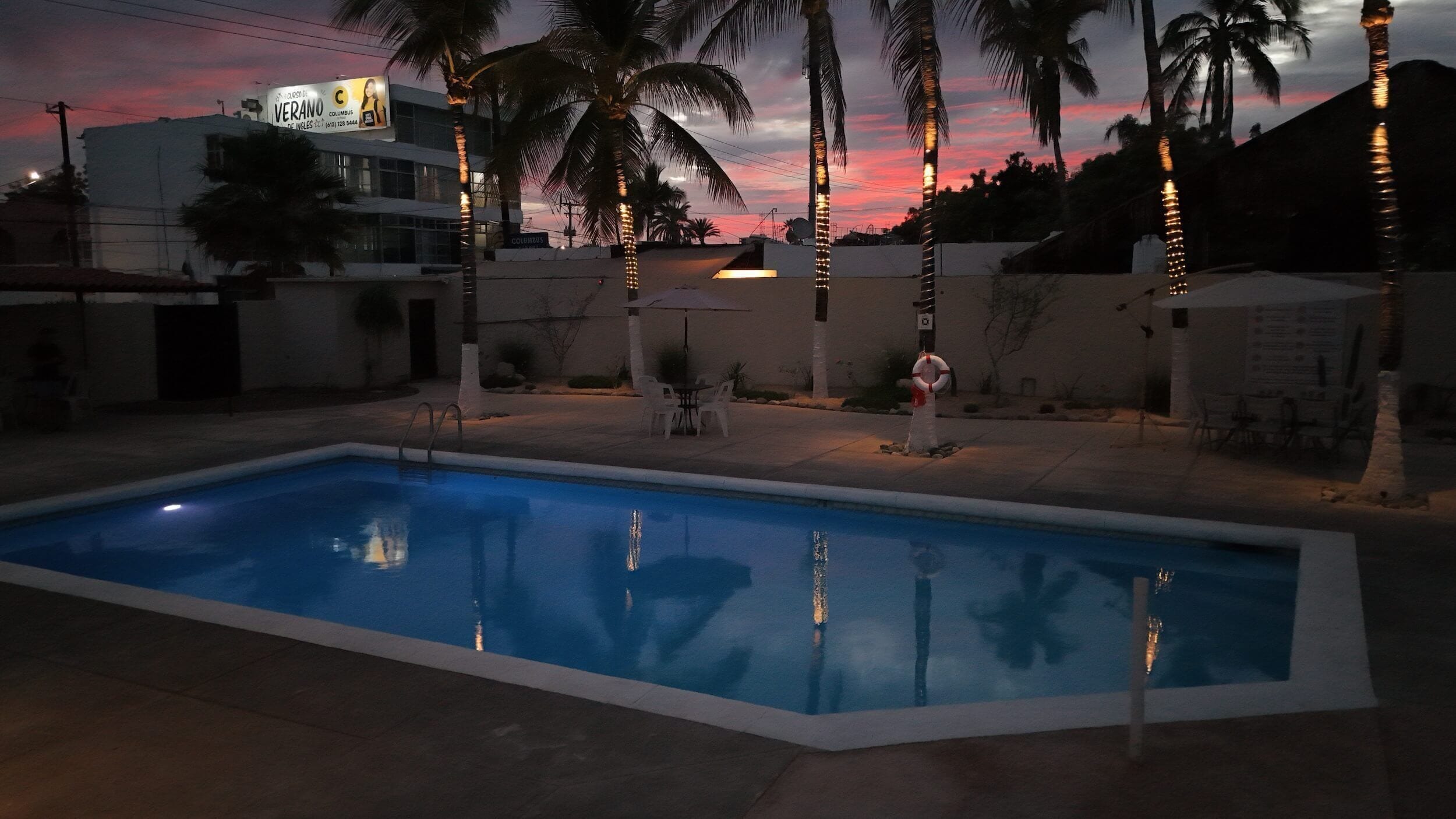 A swimming pool with a view of a building with a sign, palm trees, and a sunset.
