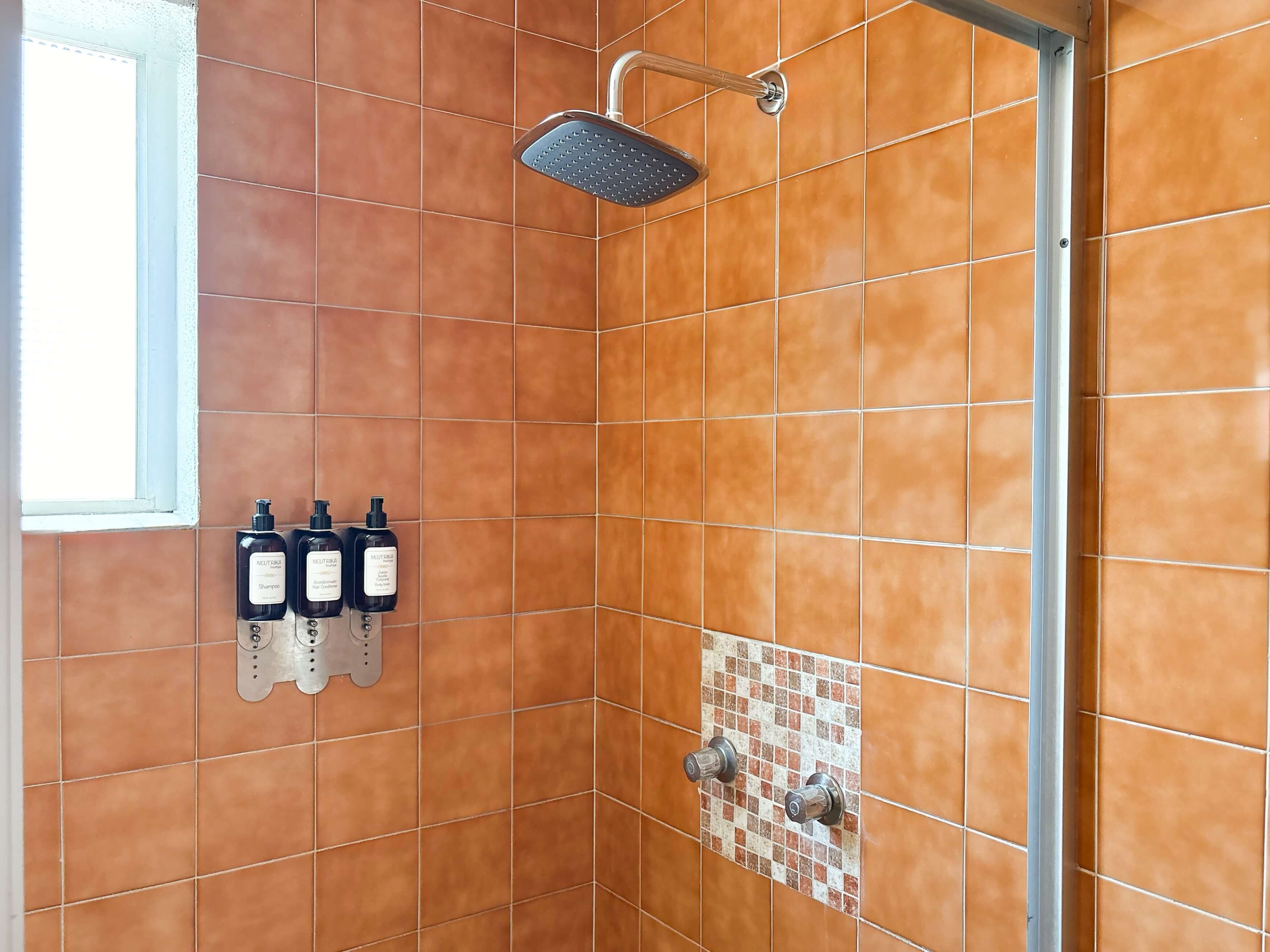 Et bruseområde med et håndholdt brusehoved, en glasdør og tre shampoo-flasker i en dispenser fastgjort til væggen.
