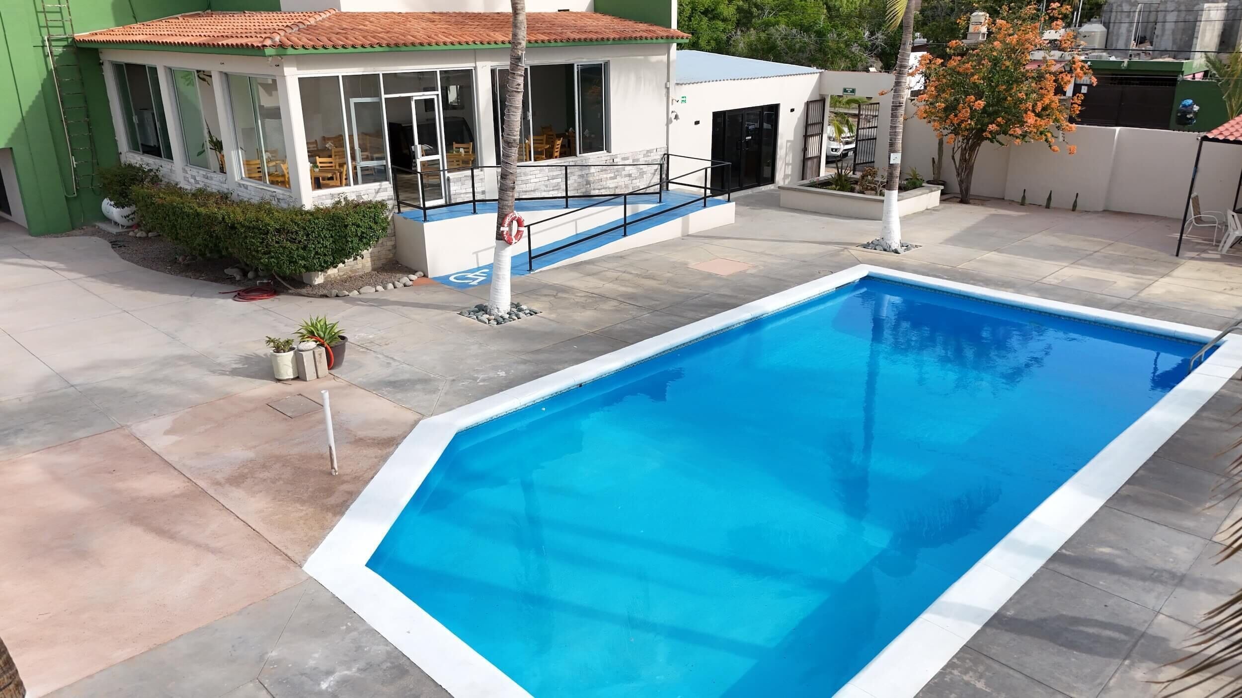 Ein Schwimmbecken mit weißem Rand, eine überdachte Terrasse mit Gartenmöbeln und ein kleines Gebäude mit einem Ziegeldach.