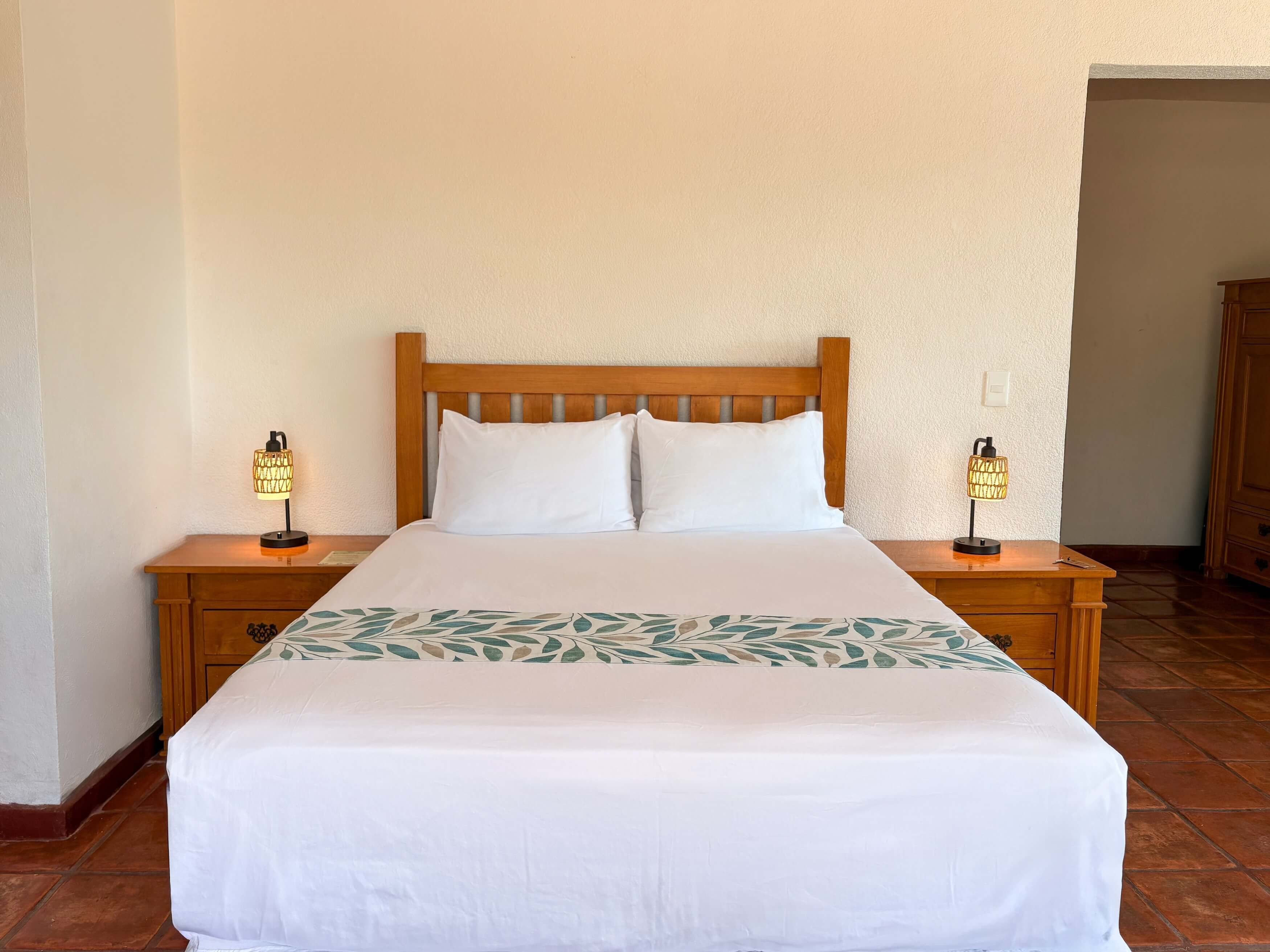Una cama bien hecha con sábanas blancas y una colcha de estampado floral, flanqueada por mesitas de noche de madera con lámparas.