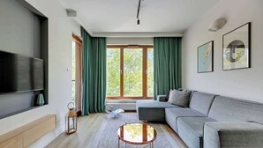A modern living room with a flat-screen TV, a grey sofa, a round coffee table, and a large window with green curtains. - Gdynia Orłowo | 450 metres to the Beach (Gdynia)