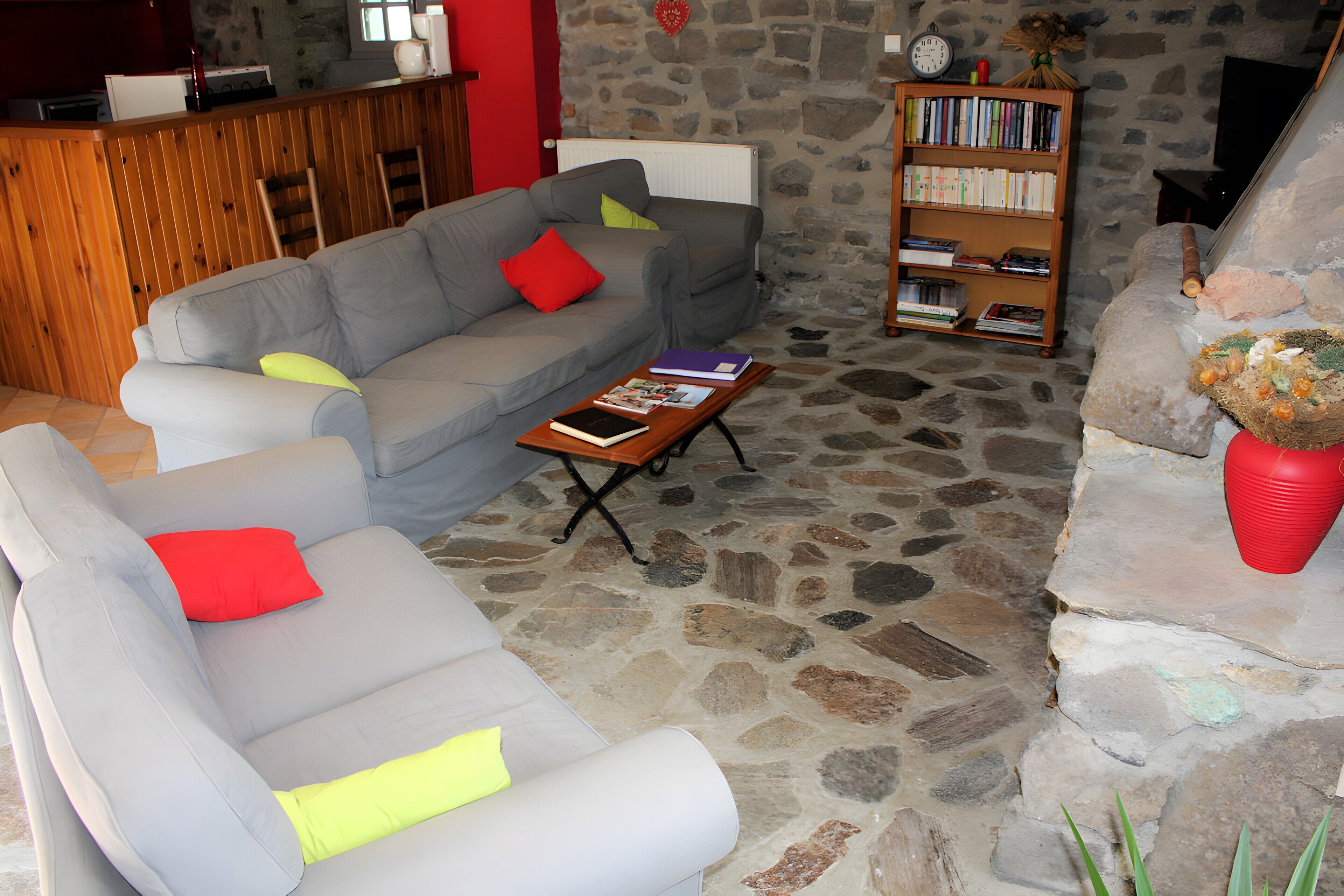 A living room with a stone floor, a grey sofa with red and yellow cushions, a wooden bookshelf, and a red vase with flowers.