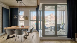 Een moderne kamer met een houten eettafel, grijs gestoffeerde stoelen, een hanglamp en een grote schuifglazen deur die leidt naar een buitenbalkon.