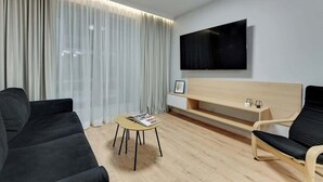 Un salon moderne comprenant un canapé, un fauteuil, une petite table, un téléviseur fixé au mur et une grande fenêtre avec des rideaux.