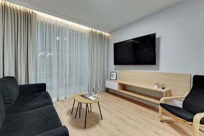 A modern living room with a sofa, a chair, a small table, a TV mounted on the wall, and a large window with curtains. - Grano Residence 2 | Riverside | Parking (Gdańsk)