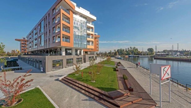 A modern multi-story building with a glass facade, a waterfront view, and a pedestrian walkway. - Brabank 17 | Riverside & Balcony | Parking (Gdańsk)