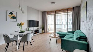 A modern living room with a green sofa, a round coffee table, a dining table with chairs, a flat-screen TV, and a large window with blinds. - Brabank 17 | Riverside & Balcony | Parking (Gdańsk)