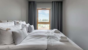 Habitació d'hotel amb un llit gran, cortines grises i vista a un paisatge urbà per la finestra.