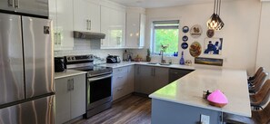 A modern kitchen with stainless steel appliances, a central island, and a window with a view of greenery. - Bright Modern Open Haven with Tree-Lined Terrace (Montréal)