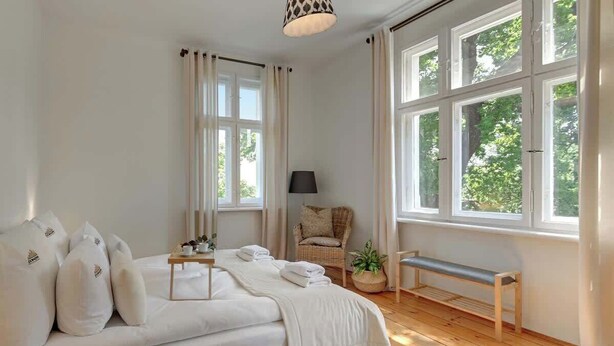 Et soveværelse med seng, bænk, stol, lampe og store vinduer.
