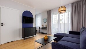 A modern living room with a sofa, a coffee table, a TV mounted on a wall, a dining area with a chair, and a large window with curtains. - Angielska Grobla 82B | Near Old Town & Marina (Gdańsk)