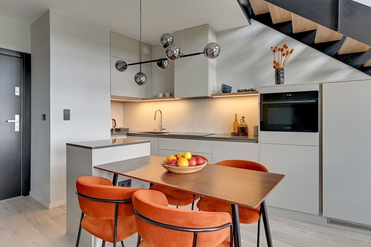 Eine moderne Küche mit einer zentralen Kochinsel, einem Essbereich mit orangefarbenen Stühlen und einer Treppe mit Holzvertäfelung.