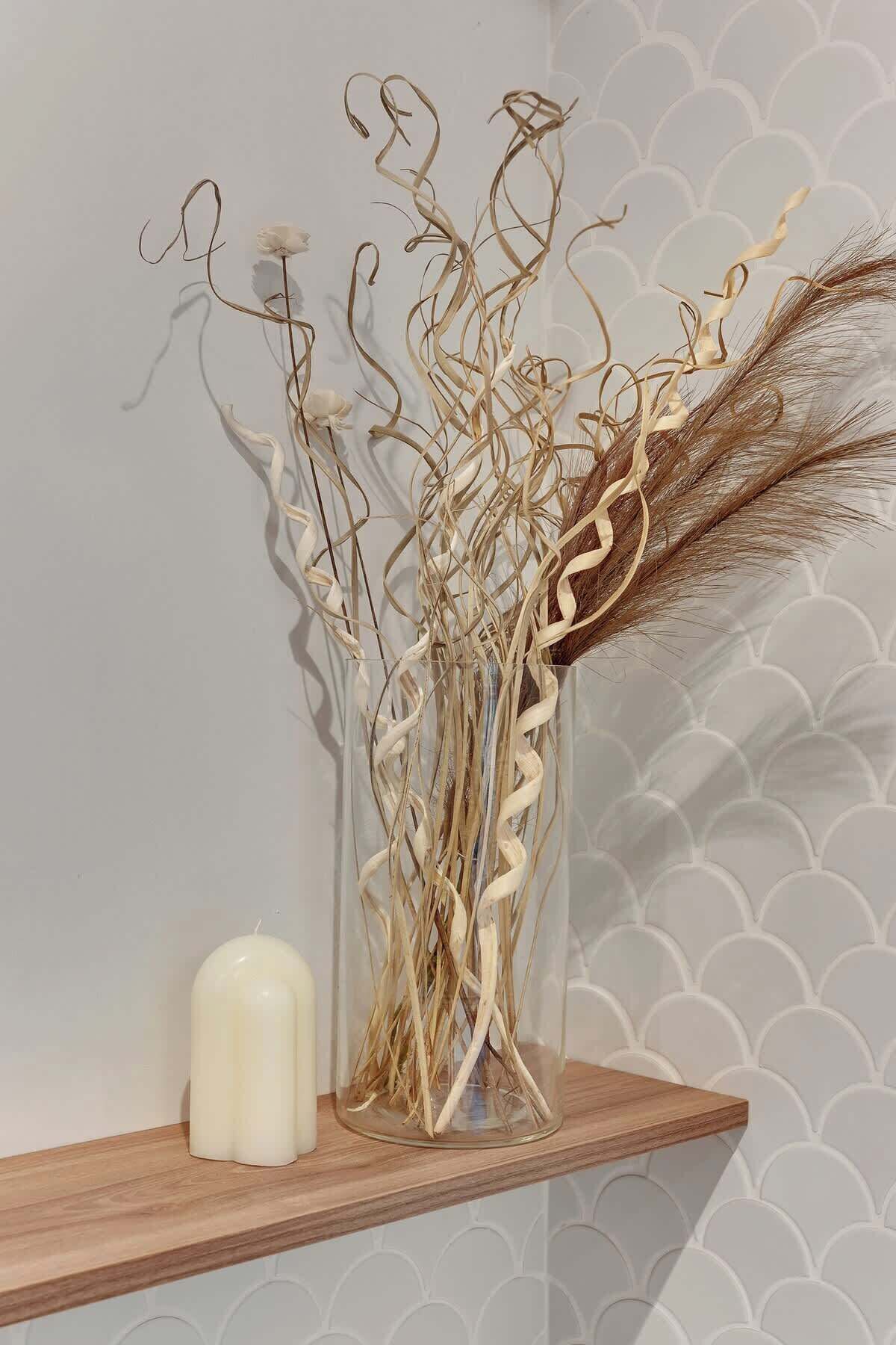 A clear glass vase with dried plants and a candle on a wooden shelf against a patterned wall.