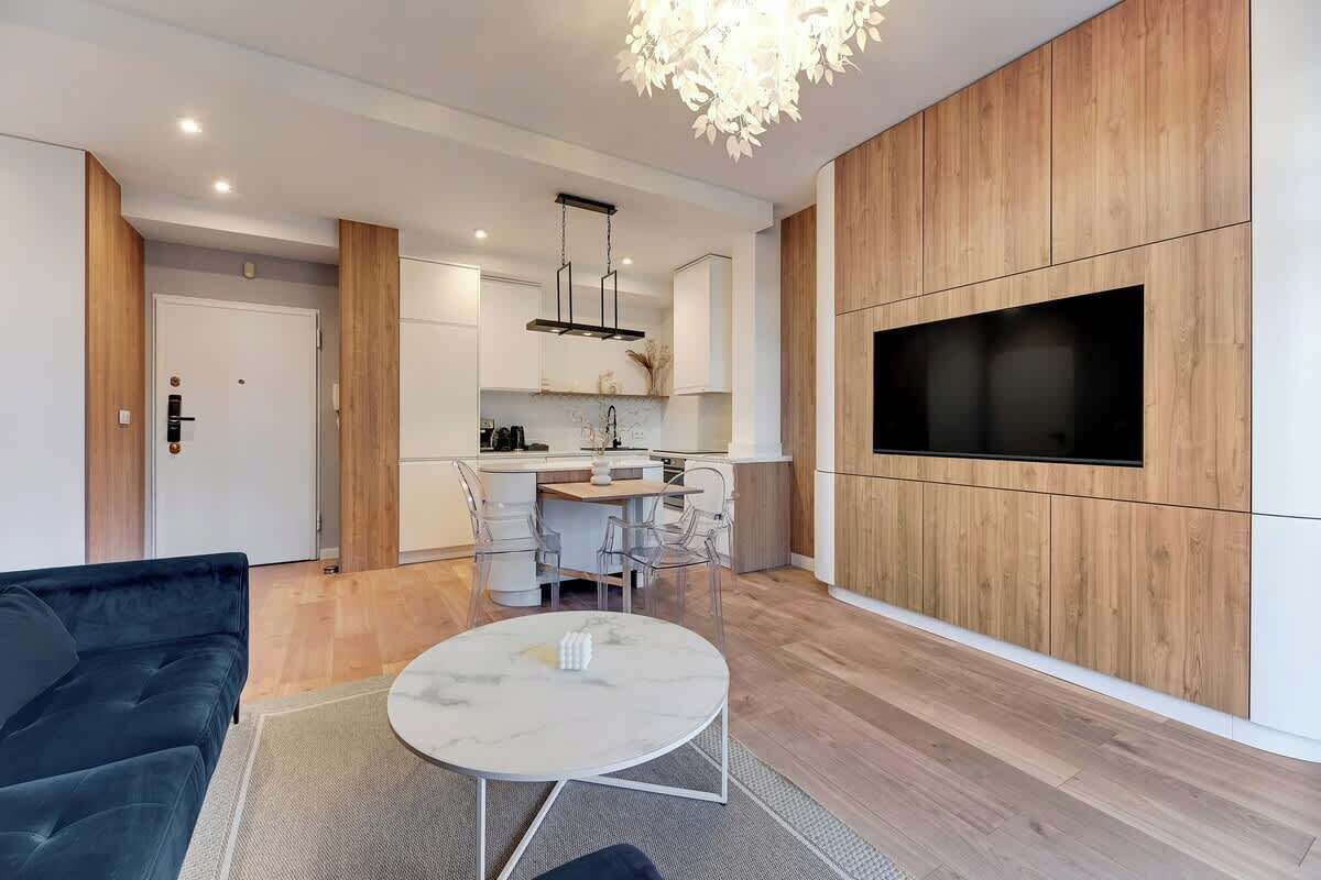 A modern living room with a sofa, a coffee table, a TV mounted on a wooden wall, and a kitchen area in the background.