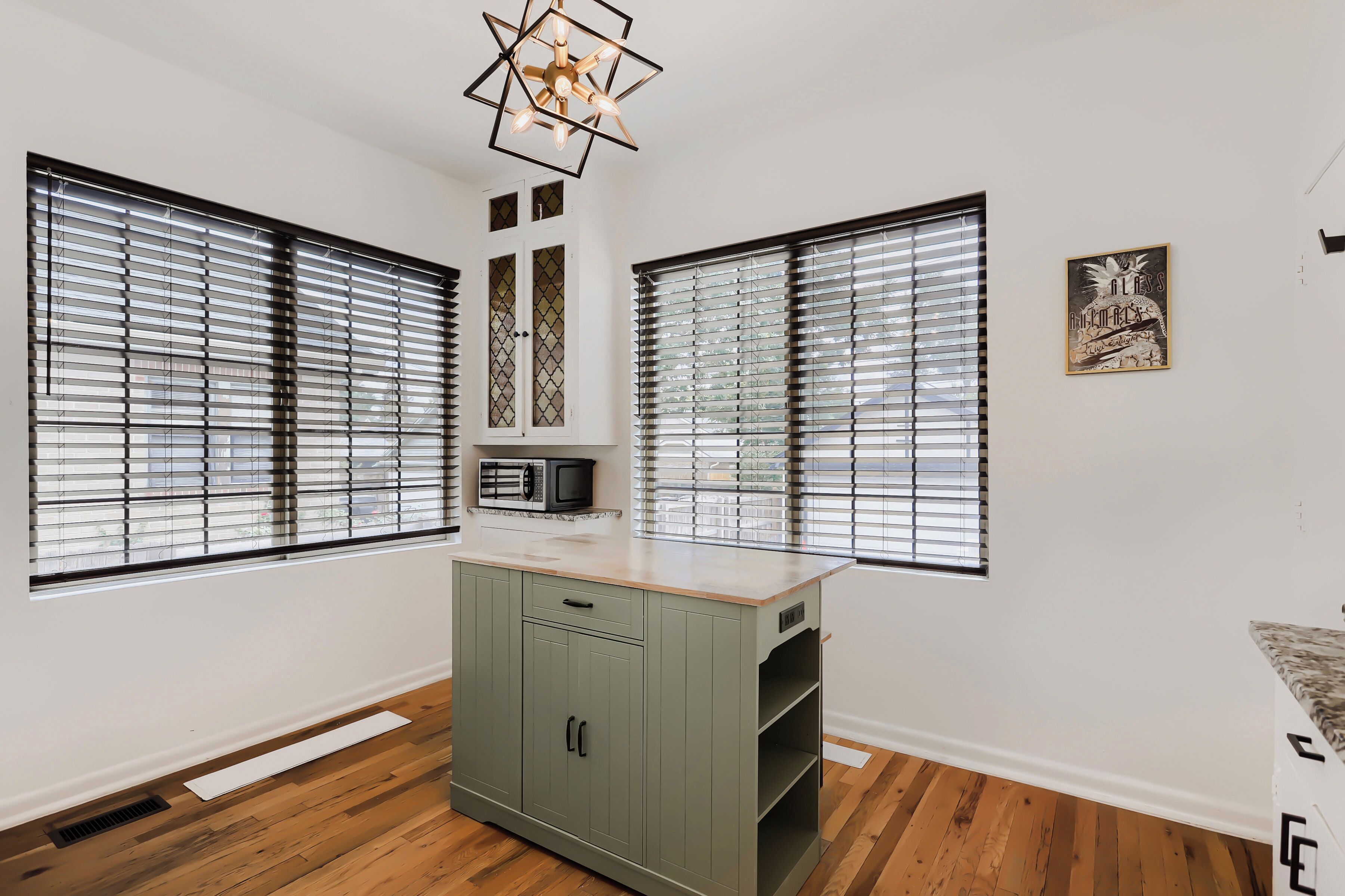 A room with two large windows with blinds, a kitchenette with a microwave, a cabinet, and a ceiling light fixture.