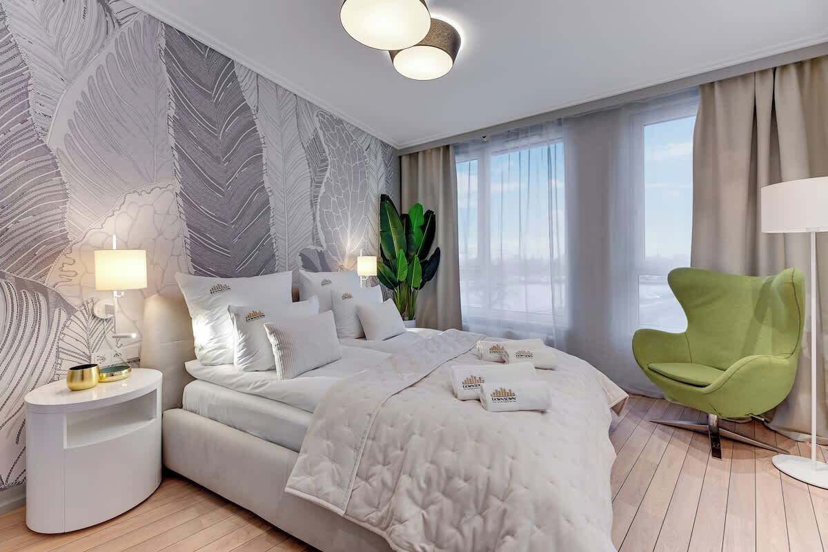 Quarto de hotel moderno com uma cama grande, uma poltrona verde, uma mesinha de cabeceira com abajur e uma grande janela com cortinas.
