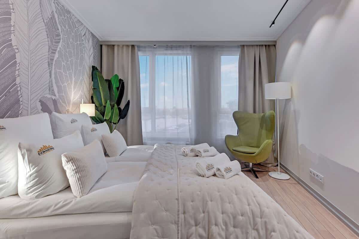 Une chambre d’hôtel avec un lit, un fauteuil vert, une grande plante et une vue sur la ville.
