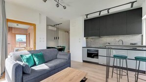 Ein modernes Wohnzimmer mit grauem Sofa, Holztisch und einer Küche mit dunklen Schränken und Barhocker.