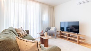 Una sala de estar con un sofá, una mesa de centro, un televisor sobre un mueble de madera y cortinas translúcidas.