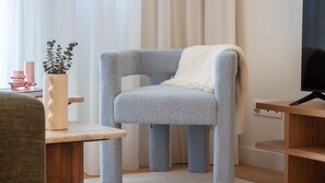 Ein Zimmer mit einem grau gepolsterten Sessel, einer weißen Tagesdecke, einem Holznachttisch, einer Topfpflanze und einem Fernseher auf einem Holzständer.