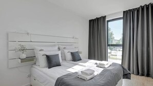 Ein modernes Schlafzimmer mit einem großen Bett, grauer Bettwäsche, einem Nachttisch mit einer Vase und einem Fenster mit Vorhängen.