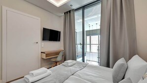 Et hotelværelse med seng, skrivebord, stol, fjernsyn og et stort vindue med gardiner.
