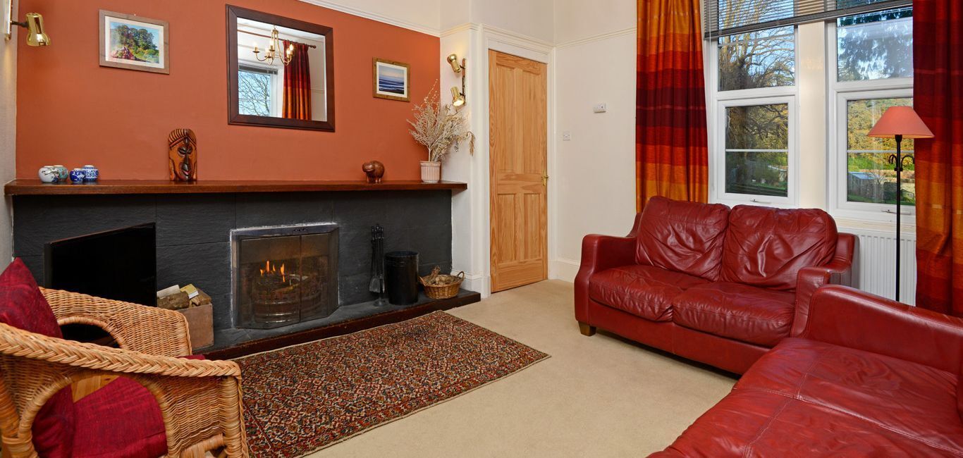 A living room with a fireplace, red leather sofa, wicker chair, and a view of the outdoors through large windows.