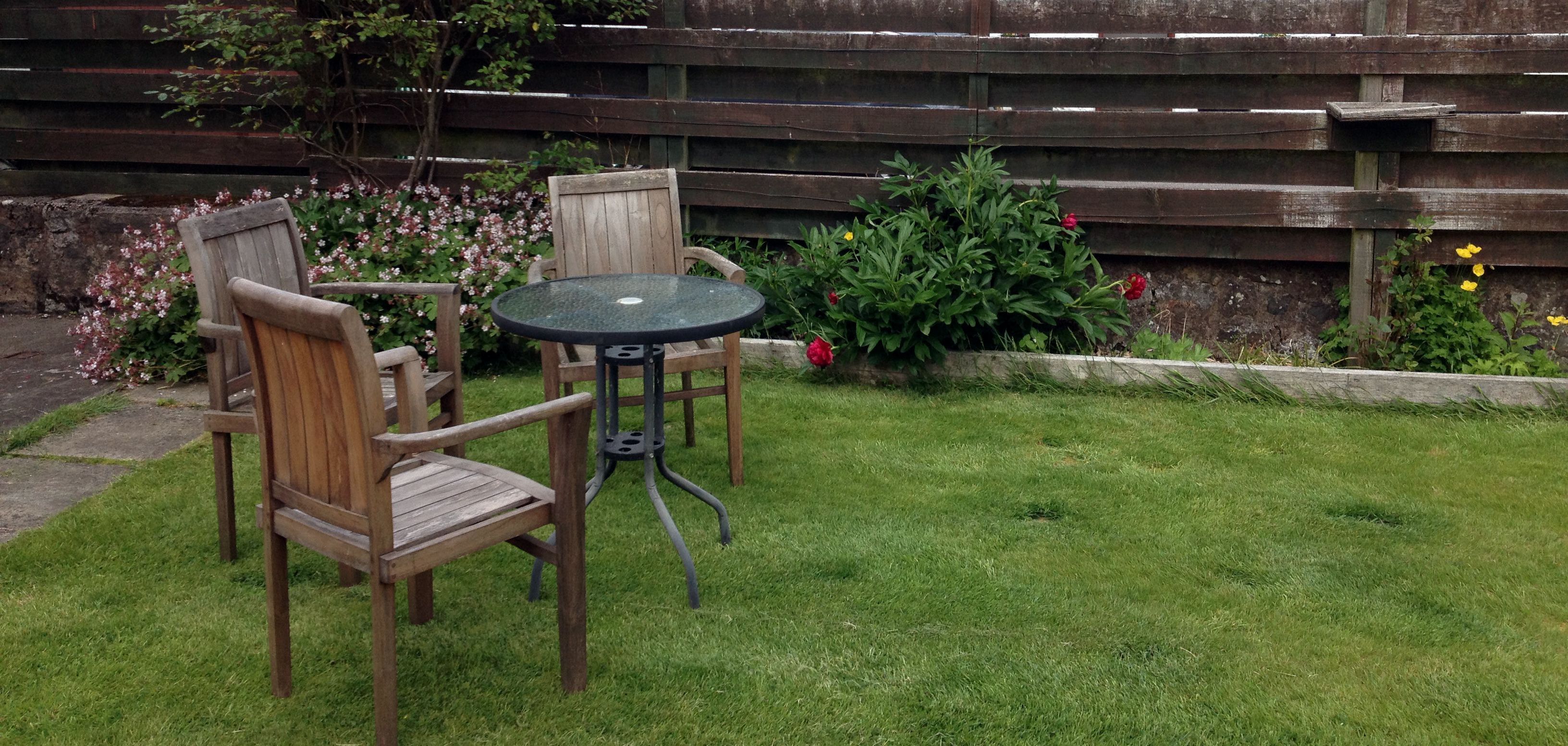 A wooden outdoor seating area with a table and chairs, surrounded by a wooden fence and flowering plants.