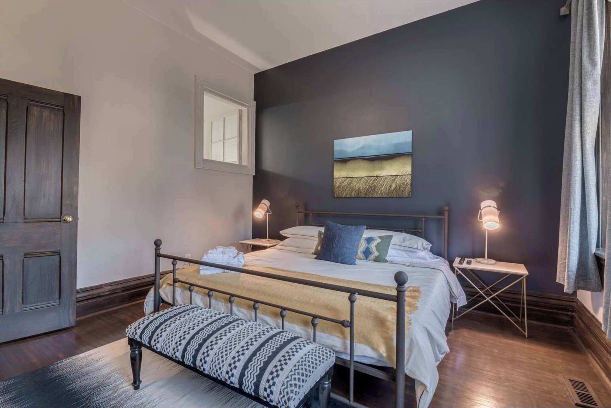 A bedroom with a bed, bedside tables, a bench, a window, and a painting on the wall.