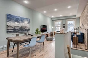 A modern living room with a wooden dining table, blue chairs, a large abstract painting, a flat-screen TV, and a staircase with a railing. - Casa Sherbourne Hotel - Modern Industrial Suite (Toronto)